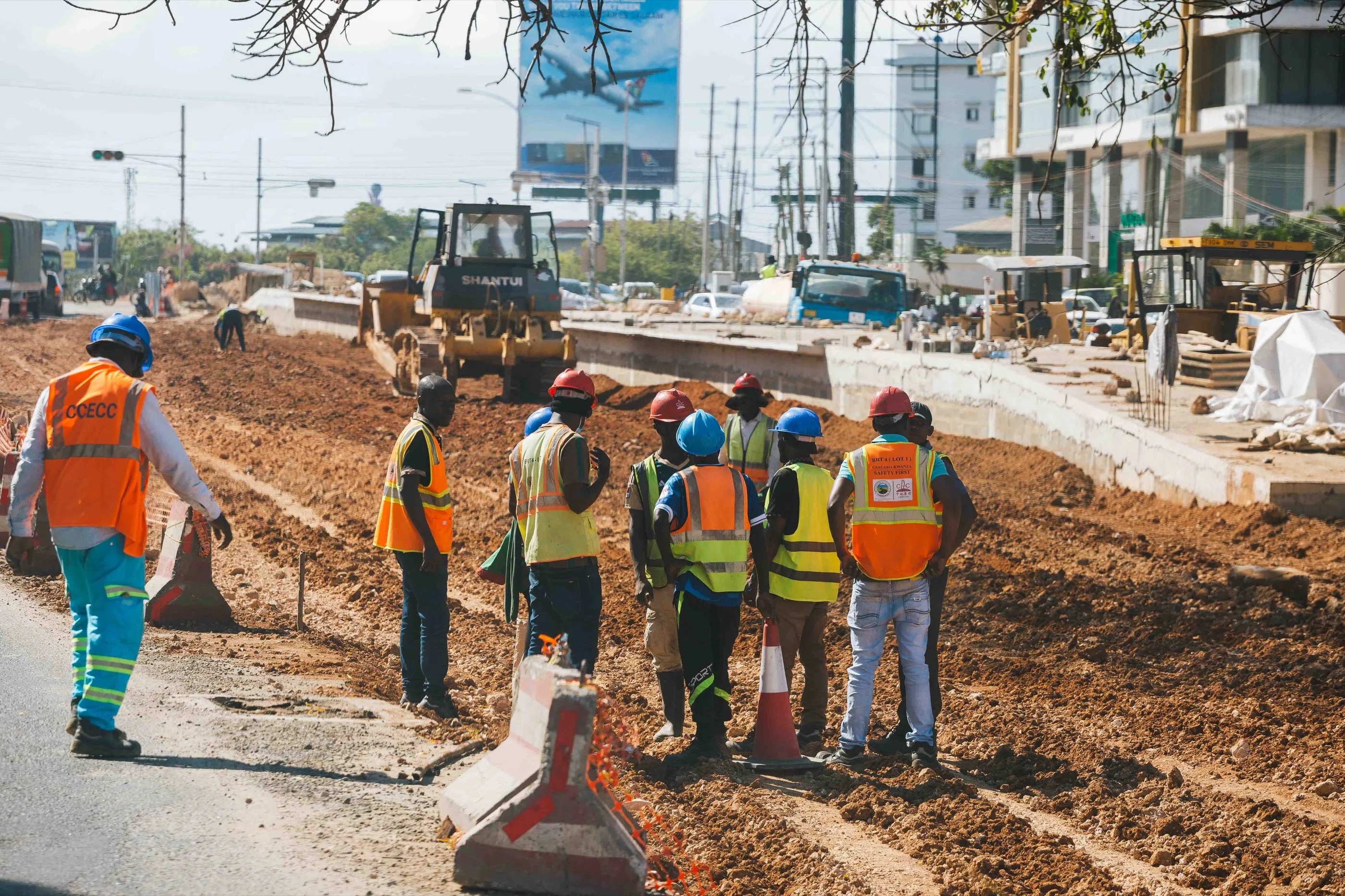 A road-construction crew in Lagos in hard hats and hi-vis vests stands in a huddle on graded red earth beside a bulldozer