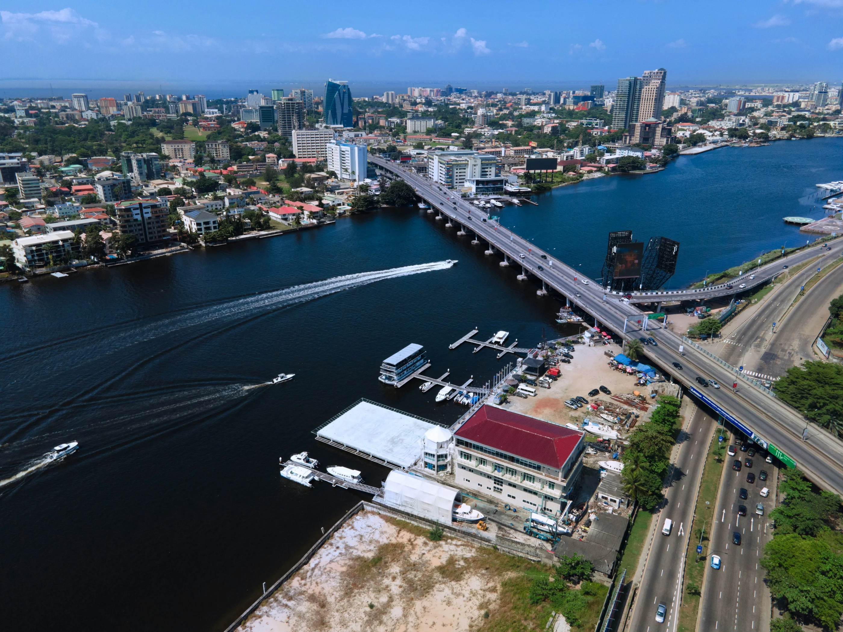 Aerial view of Victoria Island, Lagos: speedboat wakes on the lagoon, a road bridge and a waterfront of high-rise offices under a blue sky
