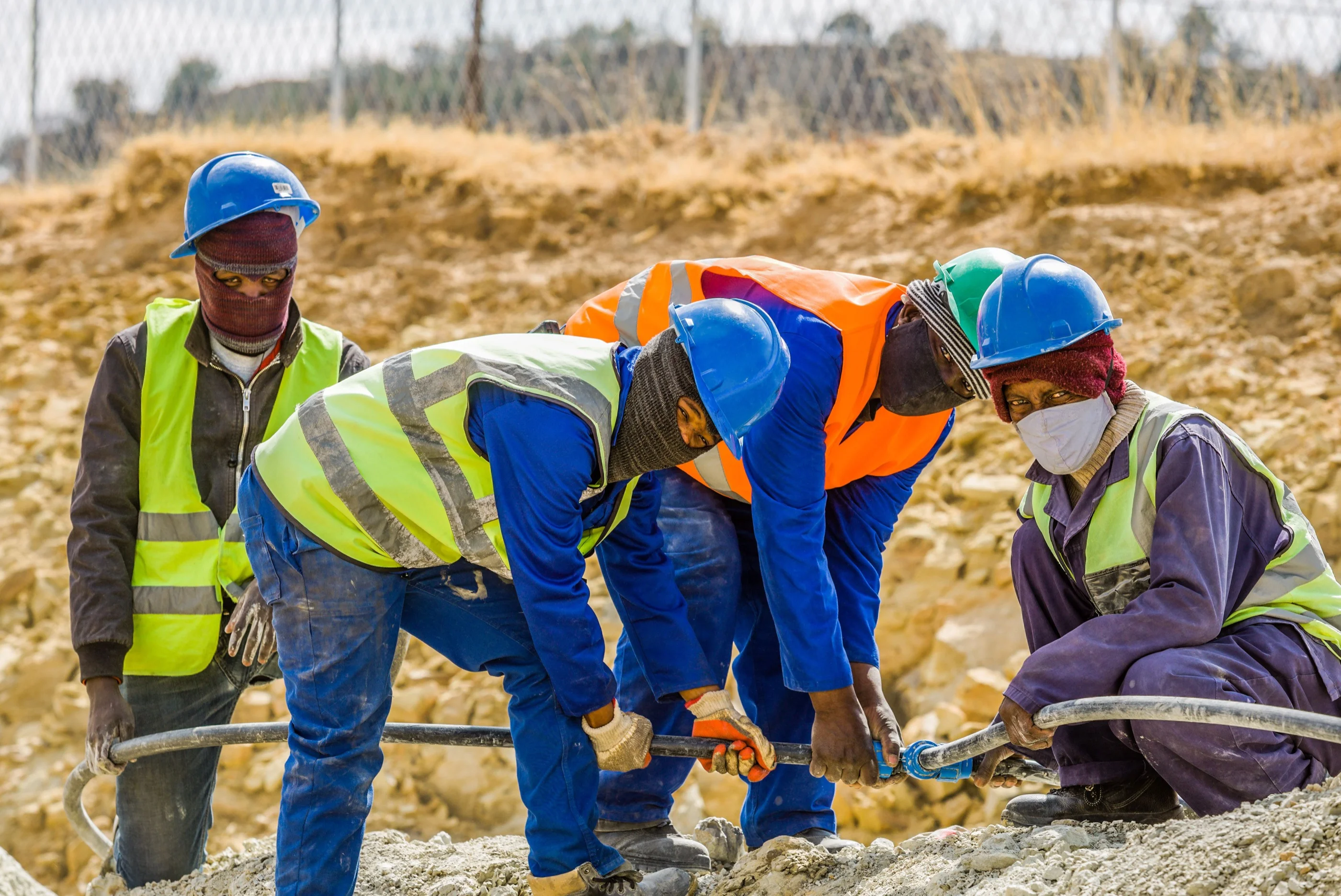 Four workers in blue hard hats and hi-vis vests bend over a hose coupling on a rocky site